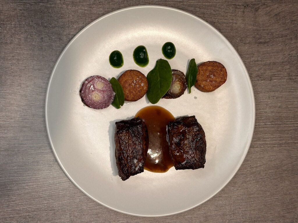 Plat gastronomique avec viande grillée, légumes rôtis, feuilles vertes et sauce sur assiette blanche