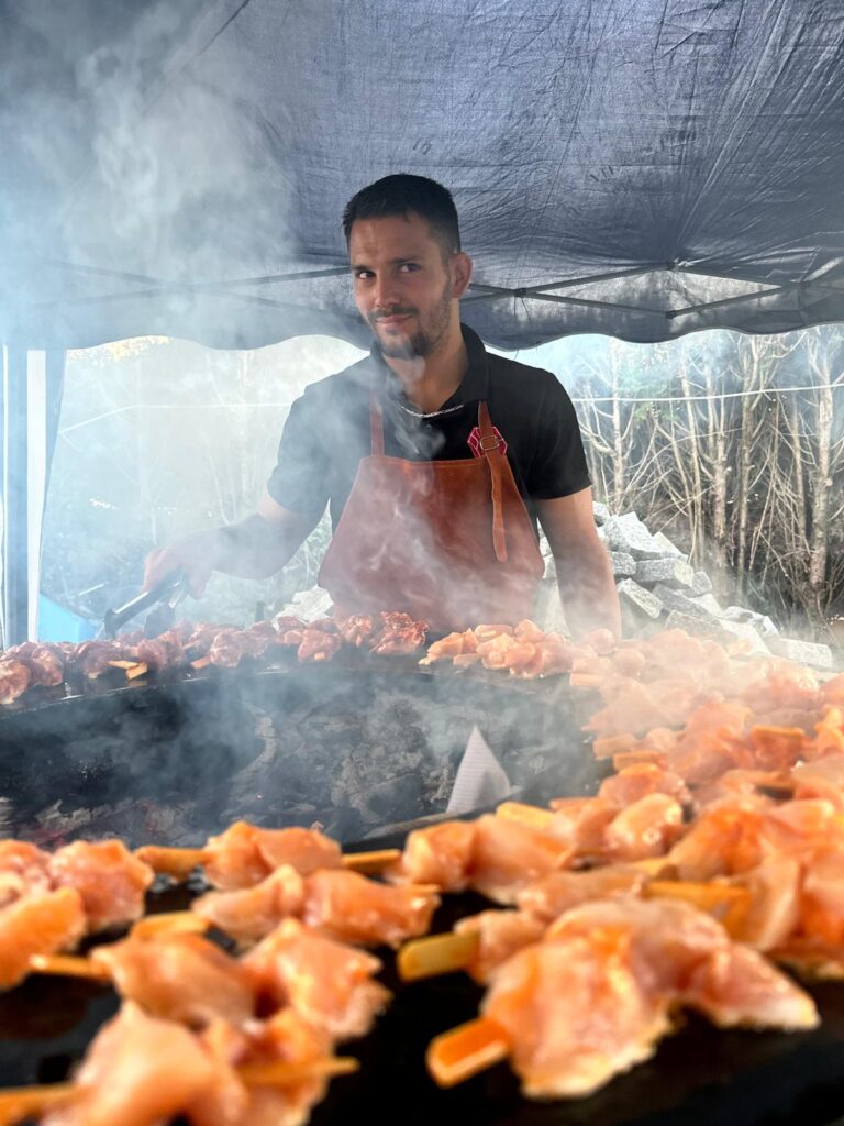 Homme cuisant des brochettes de viande sur un grand gril enfumé sous un auvent